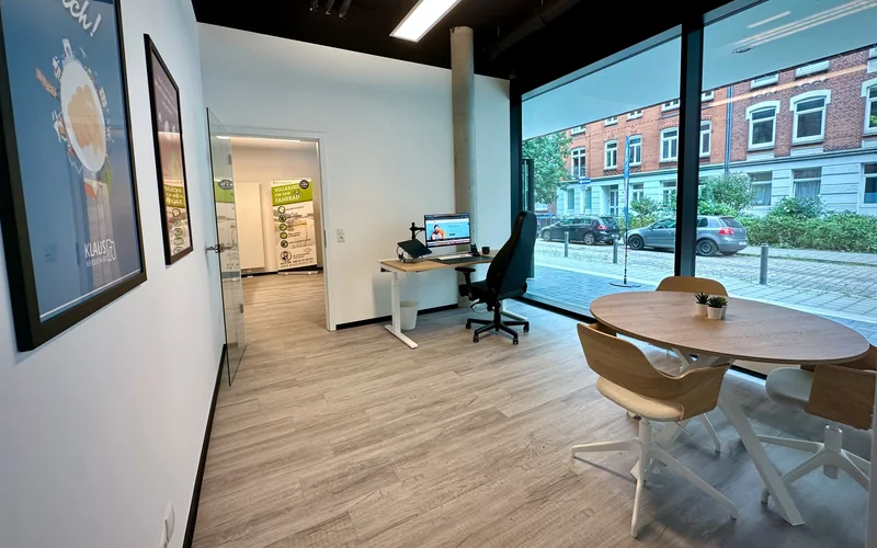 Blick in das helle Kieler Büro mit Arbeitsplatz, rundem Besprechungstisch und großer Fensterfront zur Straße